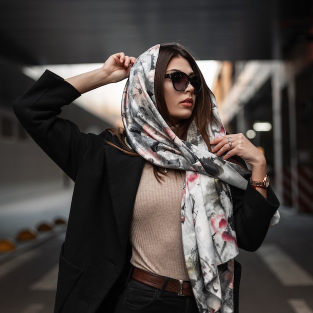 Photo elegant young pretty beautiful woman in stylish dark sunglasses in a black coat puts a luxurious silk scarf on head outdoors. beautiful girl fashion model posing on the road in the city. sexy lady.
