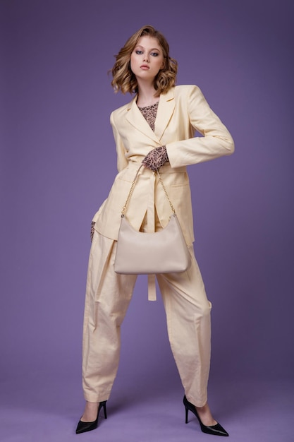 Elegant young woman in beige cream linen suit jacket trousers blazer handbad on purple background