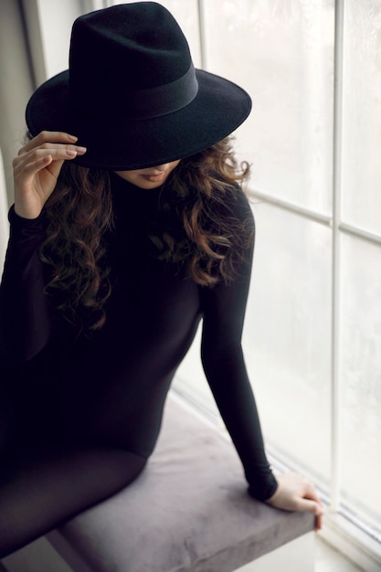 Элегантная молодая женщина брюнетка вьющиеся волосы в черной шляпе и одежде, позирует возле окна.