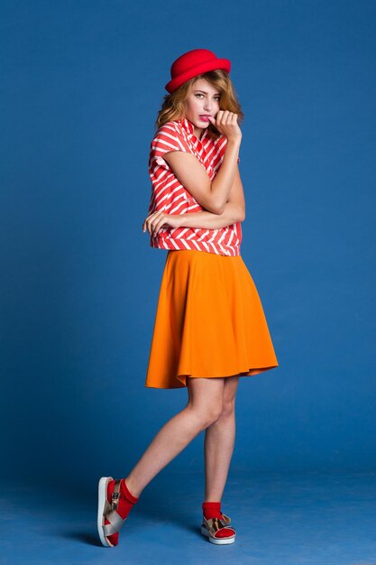 elegant young woman in orange skirt red white shirt, hat posing on blue background