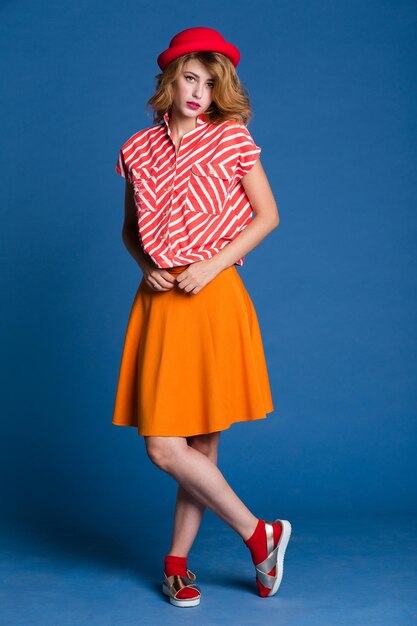 Elegant young woman in orange skirt red white shirt, hat posing\
on blue background