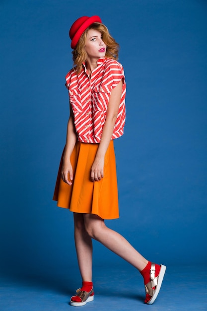 elegant young woman in orange skirt red white shirt, hat posing on blue background