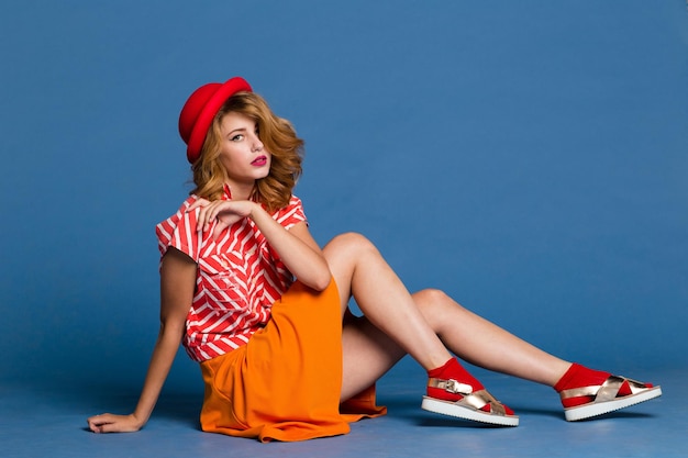 elegant young woman in orange skirt red white shirt, hat posing on blue background