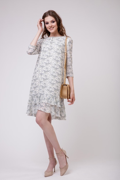 Elegant young woman in pretty beige dress with floral pattern, handbag posing on white background