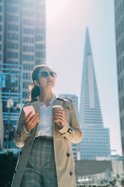 Elegant young woman standing outside of modern office building
in busy city and holding take away coffee and texting on smart
phone. beautiful lady employee in sunglasses drink morning tea in
urban