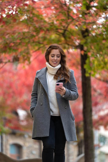 Elegant young woman walking with her phone on the street in autumn with colorful trees on the background