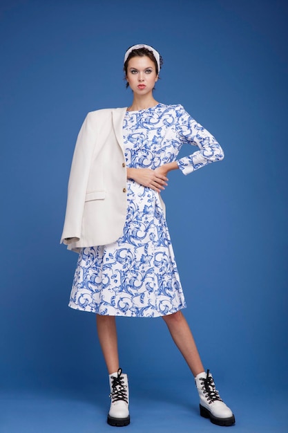 elegant young woman in white jacket, patterned dress, boots, head scarf posing over blue background