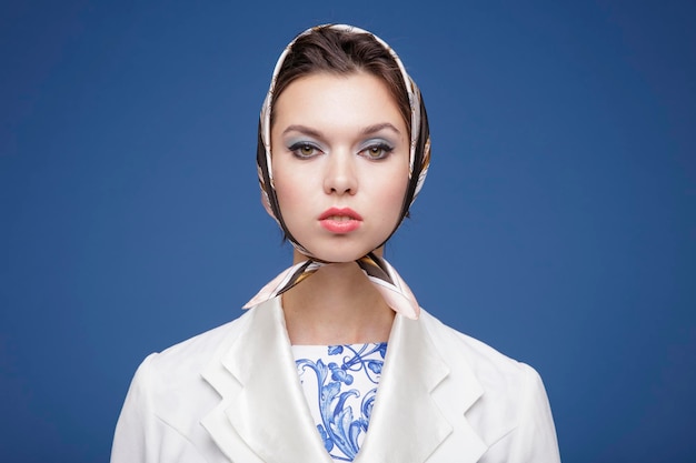 elegant young woman in white jacket, patterned dress, boots, head scarf posing over blue background