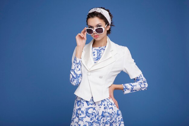 Elegant young woman in white jacket, patterned dress, boots,\
head scarf sunglasses blue background