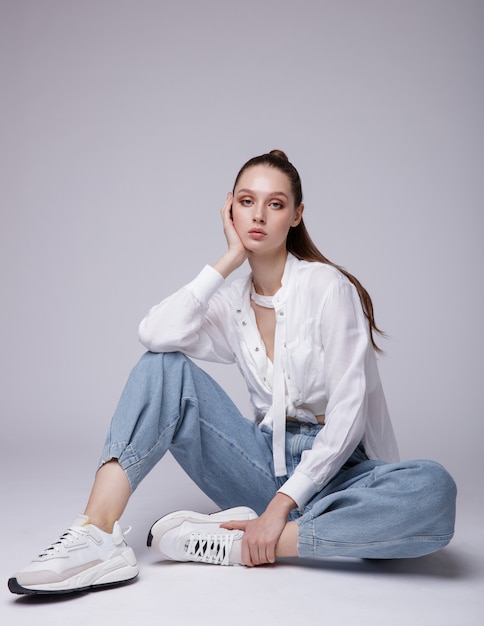 Elegant young woman in white shirt sneakers blue denim jeans on background The model is sitting