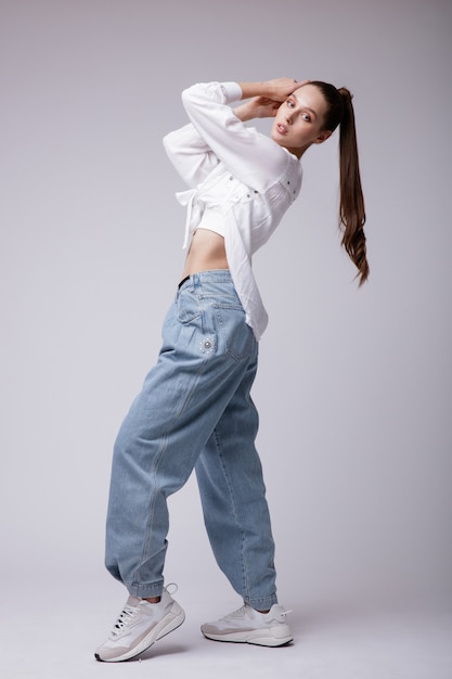 Elegante giovane donna in camicia bianca sneakers jeans blu denim su sfondo studio shot