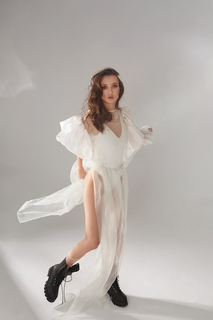 Elegant young woman with seductive body forms in a white fluttering dress and high boots high fashio...