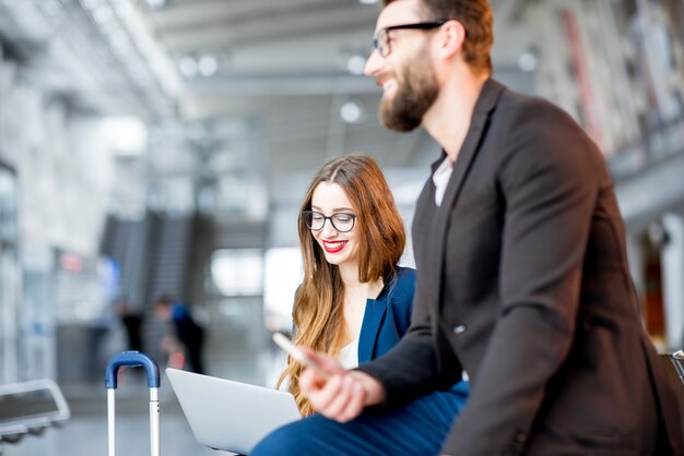 Elegant zakenpaar zit met laptop, telefoon en koffer in de wachtzaal op de luchthaven. Zakelijk reisconcept