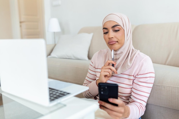 Elegante aantrekkelijke moslimvrouw die mobiele telefoon en laptop gebruikt om online winkelinformatie in de woonkamer thuis te zoeken Portret van een gelukkige vrouw die een product koopt via online winkelen