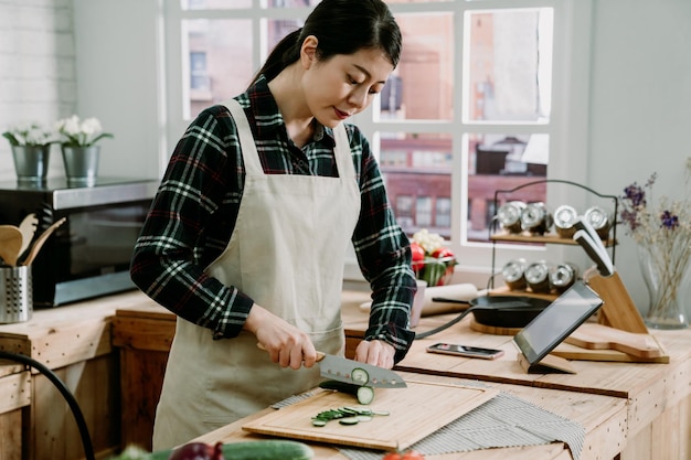 elegante aziatische chinese vrouw met een schort voor het snijden van groenten in de moderne keuken met tablet op tafel. mooie huisvrouw bereidt een gezonde maaltijd met verse komkommer in plakjes in de kookplaats binnen.