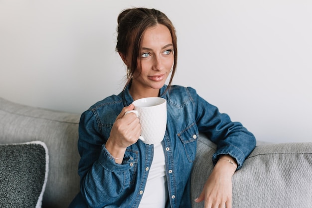 Elegante blanke vrouw met grote blauwe ogen en donker haar, gekleed in een denim shirt, zittend op de bank met koffie en kijkend naar het raam met een gelukkige glimlach over geïsoleerde achtergrond