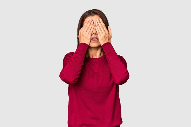 Elegante blanke vrouw van middelbare leeftijd in de studio die bang is en haar ogen met haar handen bedekt.