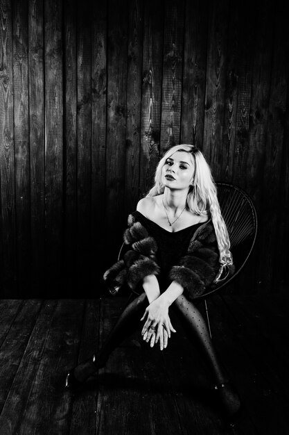 Photo elegante blonde girl wear on fur coat sitting on chair at studio against wooden background.