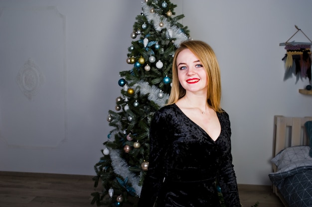 Elegante blonde meisjesslijtage op zwarte kleding tegen nieuwe jaarboom met kerstmisdecoratie.