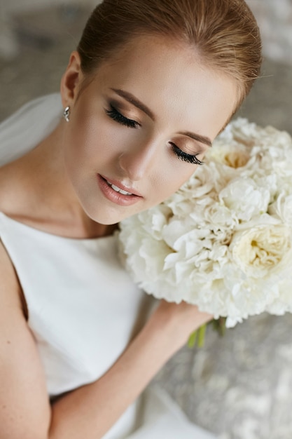 Foto elegante blonde model meisje met lichte bruiloft make-up poseren met gesloten ogen en boeket bloemen