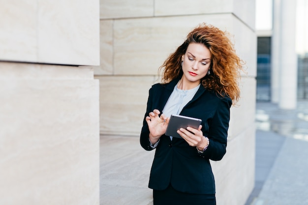 Elegante dame met krullend haar, het dragen van zwart pak, het typen van berichten of het maken van bedrijfsrapporten tijdens het gebruik van tabletcomputer