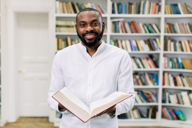 Elegante donkere mannelijke student in wit overhemd leesboek terwijl je in de bibliotheek tegen boekenplanken.