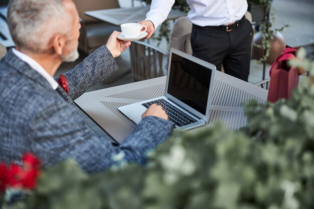 Elegante grijsharige man die een kopje koffie krijgt van een ober terwijl hij op de laptop in café werkt