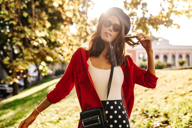 Elegante huid meisje met golvend donker haar dragen stijlvolle baret en zonnebril witte top rood shirt poseren tegen de achtergrond van zonnige herfst stad en kijken naar de camera