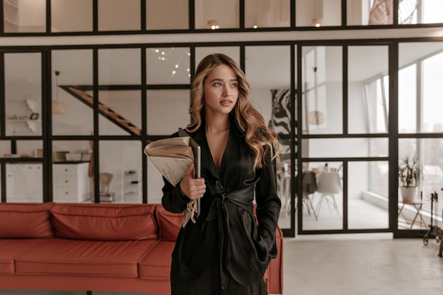 Elegante jonge blonde krullende vrouw in een stijlvol zwart zijden pak kijkt weg in de woonkamer en ho