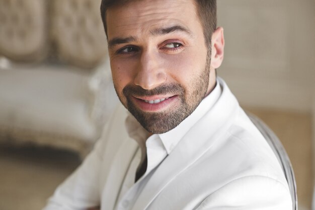 Elegante jonge knappe man met een baard in een wit klassiek pak. Het interieur van het hotel. Fotostudio. Bank. Raam. Stoel. Kroonluchter