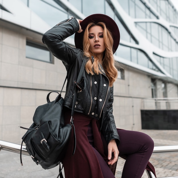 Elegante jonge vrouw met rood haar in een zwarte jas met een leren rugzak in trendy broek