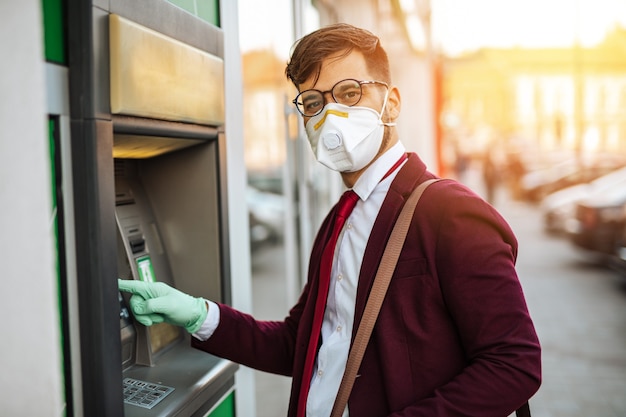 Elegante jongeman met beschermend masker die op straat in de stad staat en ATM-machine gebruikt met beschermende handschoenen op handen. Virus pandemie preventie en gezondheidszorg concept.