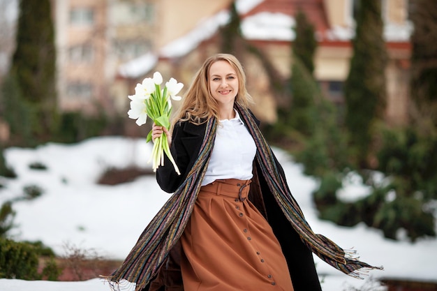 Elegante lachende jonge vrouw met lang blond haar in zwarte jas in de stad Vrouw met bloemen