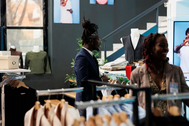 Elegante man kijkt naar trendy kleding op hangers in de showroom, winkelend voor modieuze koopwaar om de garderobe te vergroten. Afro-Amerikaanse klant koopt nieuwe modecollectie in moderne boetiek