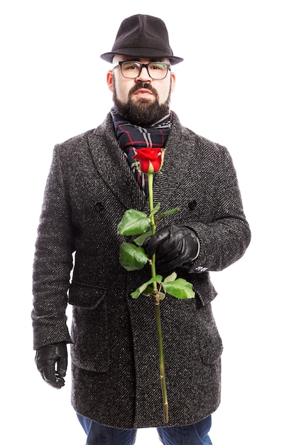 Elegante man met een baard, gekleed in een stijlvolle jas en hoed met een rode roos in zijn handen. Geïsoleerd via witte muur. Verticaal.