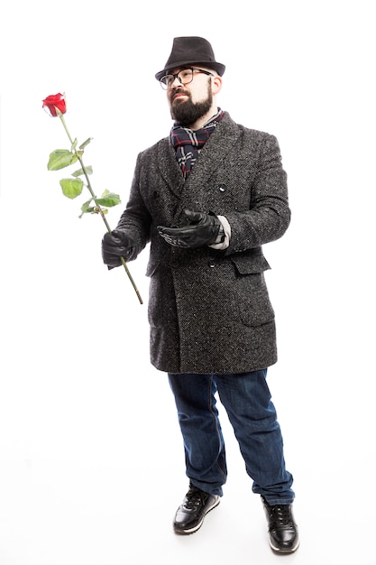 Elegante man met een baard, gekleed in een stijlvolle jas en hoed met een rode roos in zijn handen. Volledige hoogte. Geïsoleerd via witte muur. Verticaal.