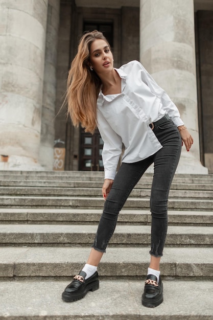 Elegante mode schoonheid vrouw model met stijlvolle casual outfit met wit overhemd jeans en schoenen staat op trappen in de buurt van een vintage gebouw in de europese stad