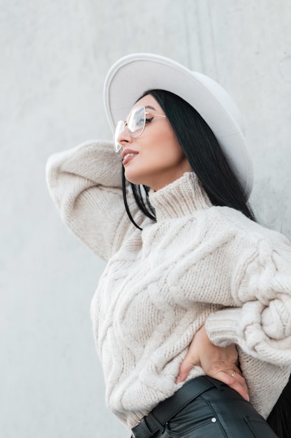 Elegante mooie jonge vrouw met een mooie bril en een hoed in een vintage witte gebreide trui staat in de buurt van een betonnen grijze muur