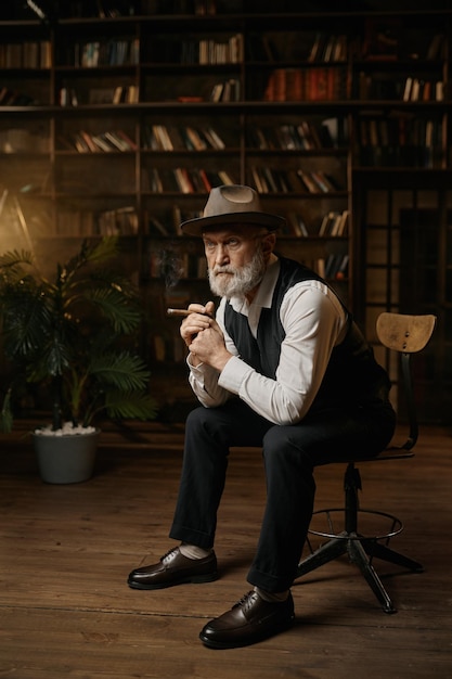 Elegante oudere zakenman of maffiabaas zittend op een vintage stoel die sigaar binnen rookt terwijl hij rust in de thuisbibliotheek of kast