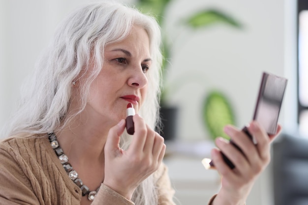 Elegante rijpe vrouw met lang grijs haar past rode lippenstift toe kijkend naar aantrekkelijke spiegel