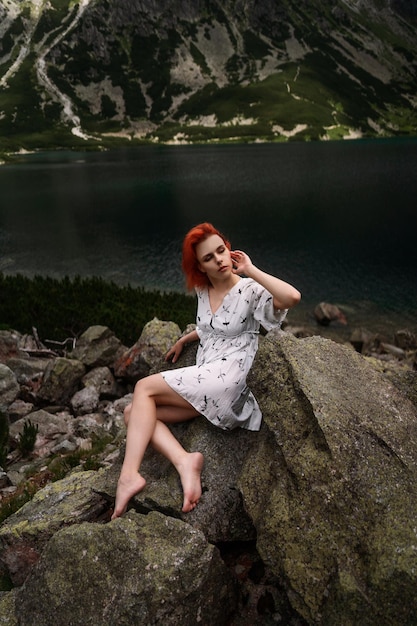 Elegante sexy dame met rood haar in een witte jurk zit op de steen naast het meer in de bergen