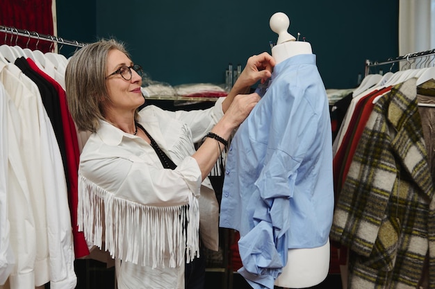 Elegante stijlvolle rijpe vrouw naaister werkt met een blauw handgemaakt shirt en probeert het op een naai-etalagepop in haar studio voor modeontwerp en kleermakers