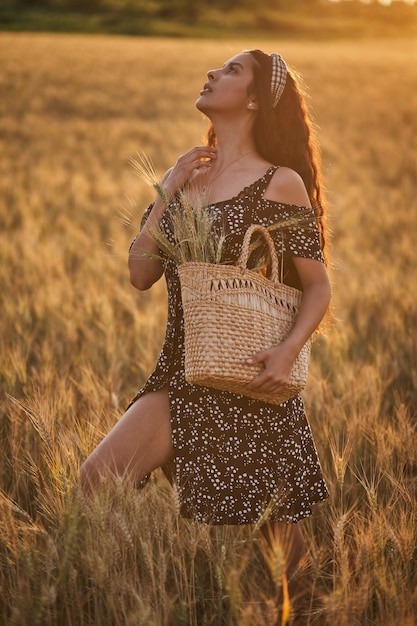 Elegante vrouw in een jurk met mand in het tarweveld bij zonsondergang.