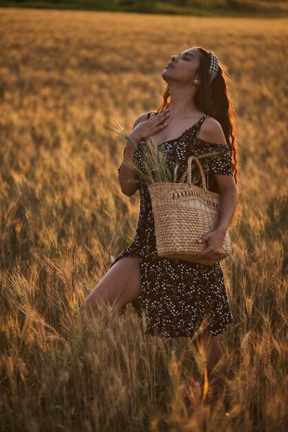 Elegante vrouw in een jurk met mand in het tarweveld bij zonsondergang.