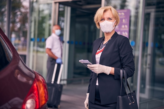 Elegante vrouw met beschermend gezichtsmasker met vliegticket en paspoort terwijl meneer reiskoffer draagt