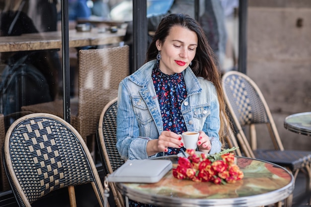 Elegante vrouw met lang donkerbruin haar in een blauwe jurk zit alleen met bloemen in het straatcafé en koffie drinken