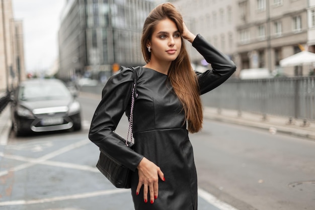 Elegante zakelijke mooie vrouw model in stijlvolle zwarte leren jurk met zwarte modieuze tas wandelingen in de stad in de buurt van een weg
