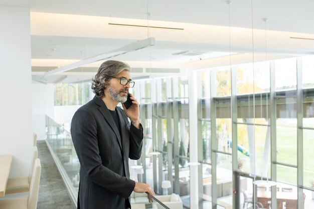 Elegante zakenman in brillen en formalwear die klant of collega telefonisch raadpleegt of een afspraak maakt in het zakencentrum