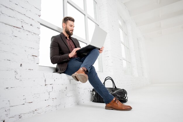 Elegante zakenman zittend op het raam met laptop in het witte kantoorinterieur