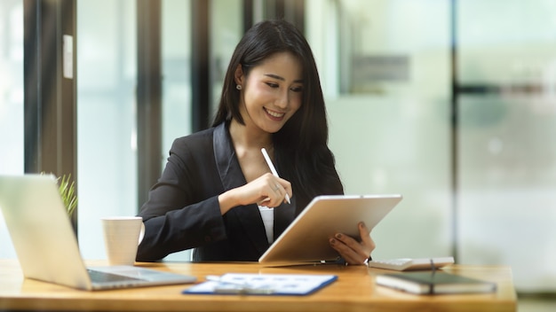 elegante zakenvrouw in zwart pak glimlacht terwijl ze naar het financiële rapport kijkt via tablet en pen vasthoudt